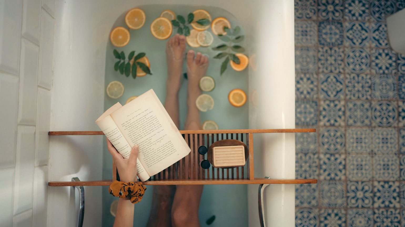 A Woman relaxing and reading in a bath with orange slices, essential oils and beautiful bathroom tiles